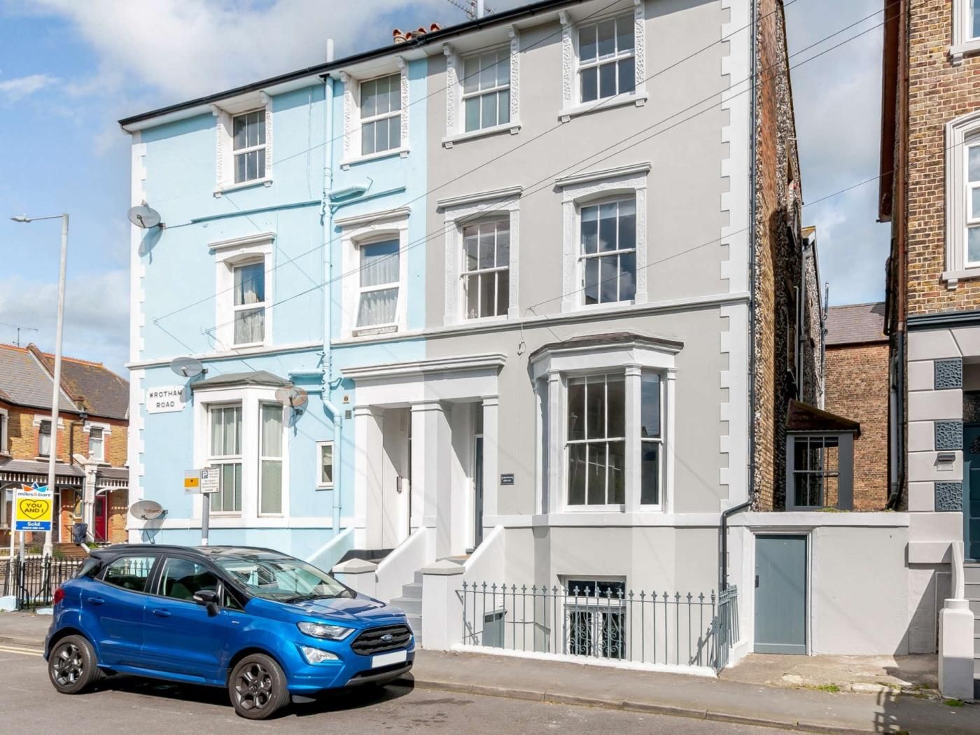 A two-story residential building with a blue and white facade, a front entrance with a staircase, and a parked blue car on the street.