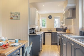 A modern kitchen with a dishwasher, stove, and sink. There is a dining table with chairs and a clock on the wall. - 3 Bed in Dawlish (oc-72972) (Dawlish)