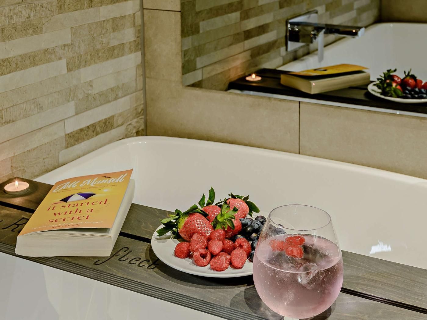 Een badkamer met een badkuip, een boek met de titel 'It Started with a Secret', een bord met aardbeien en frambozen, en een glas roze drank.