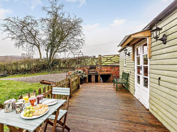 A rustic outdoor patio with a wooden table set for breakfast, a brick oven, and a wooden fence. - 1 Bed in Leighton (oc-80727) (Leighton)
