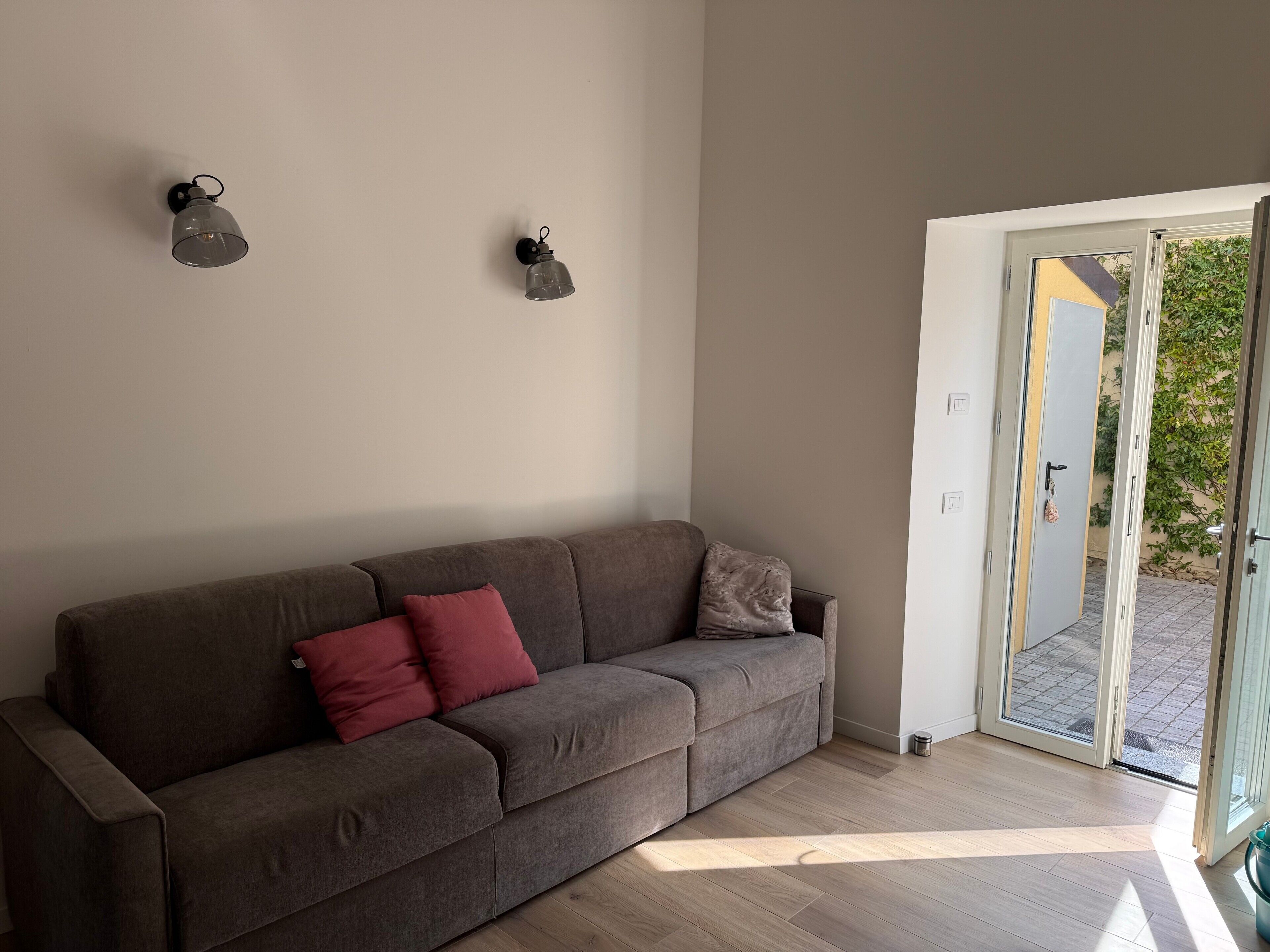Quarto em tons neutros, com um sofá cinzento, duas luminárias fixadas na parede e uma porta de vidro deslizante para o exterior.
