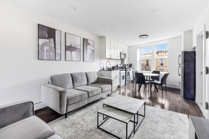 A modern living room with a grey sofa, a coffee table, and a dining area with a table and chairs. There is a kitchen area with cabinets and a microwave. - Charming NYC Two Bed! (Ridgewood)