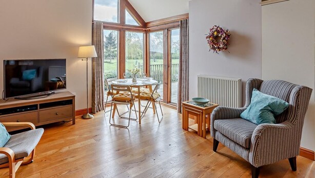 Ein Wohnzimmer mit Fernseher, einem Esstisch mit Stühlen, einem gestreiften Sessel und Blick durch große Fenster.