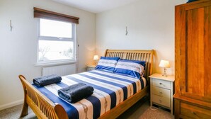 Un dormitorio con cama de madera, una ventana, un radiador, una mesita de noche con lámpara y un armario.