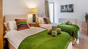 Quarto de hotel com duas camas, uma mesinha de cabeceira, um abajur, um radiador, uma janela com cortinas e um quadro na parede.