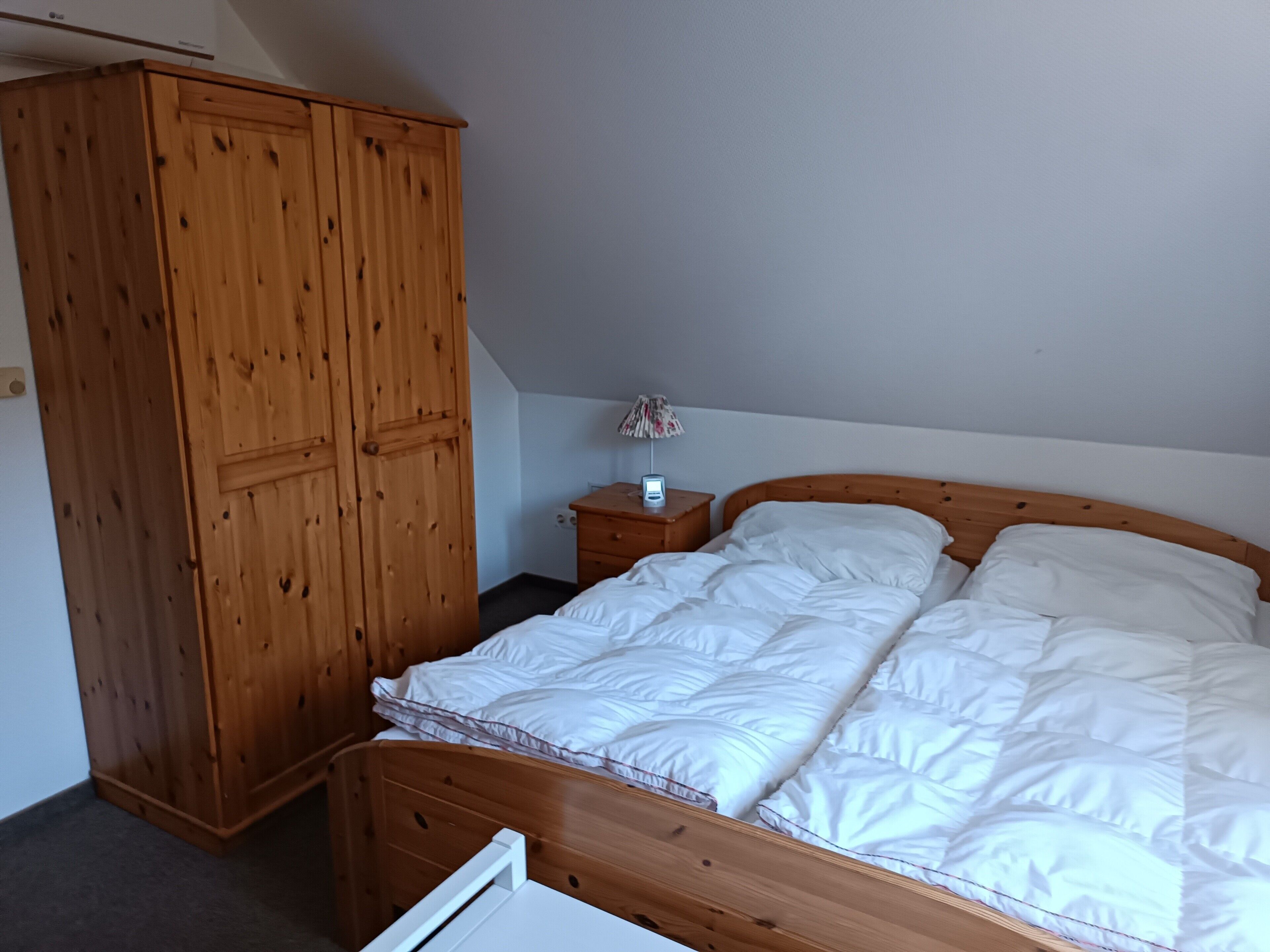 A bedroom with a wooden wardrobe, a bed with white bedding, a nightstand with a lamp, and a small table.