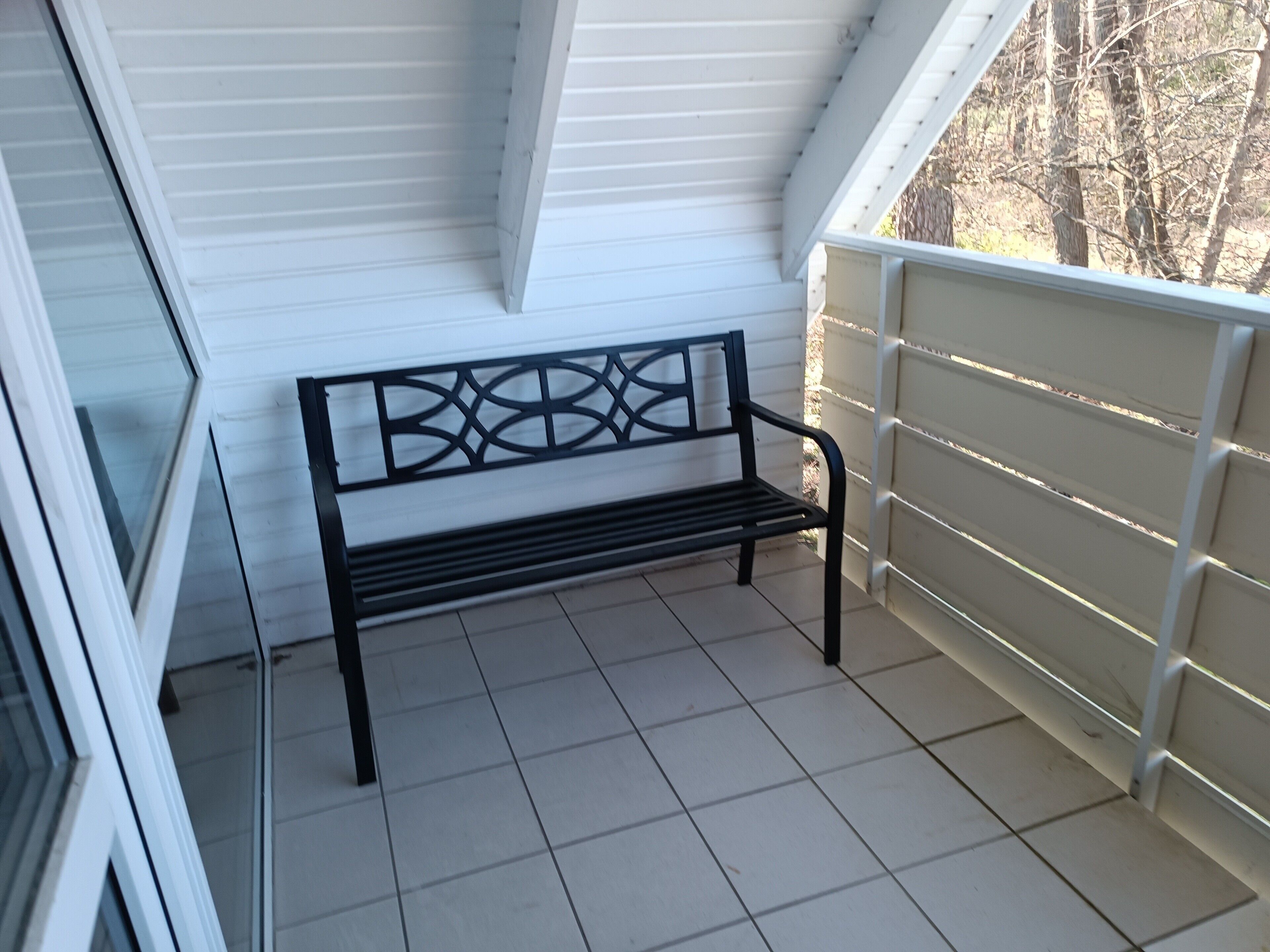 Eine schwarze Metallbank auf einem mit Fliesen belegten Balkon mit weißen Wänden und einem weißen Geländer.