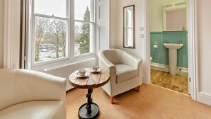 Una habitación con un gran ventanal, un sillón blanco, una pequeña mesa redonda con vajilla, y un baño visible a través de una puerta abierta.