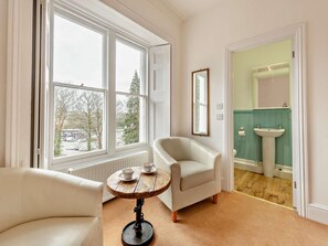 A room with a large window, a white armchair, a small round table with dishes, and a bathroom visible through an open door. - 3 Bed in Tenby (oc-73606) (Tenby)