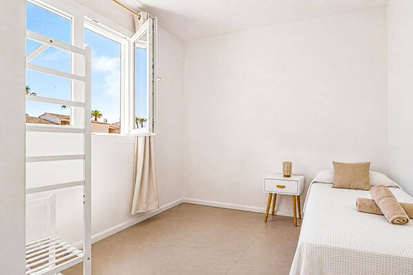 A bedroom with a bed, a nightstand, a window with curtains, and a view of palm trees.