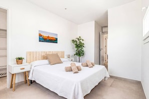 Ein Schlafzimmer mit Bett, Nachttisch, einem an der Wand hängenden Bild, einer Topfpflanze und einer Tür zu einem anderen Raum.
