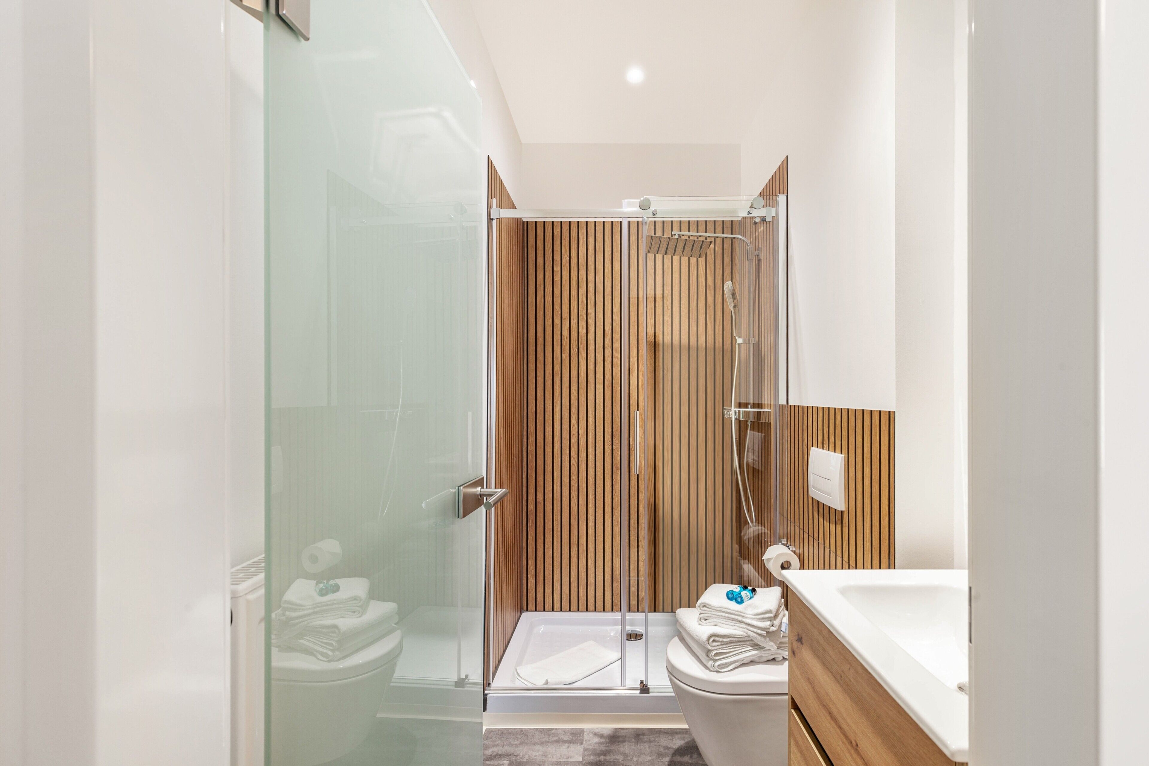 Ein modernes Badezimmer mit einer Duschkabine aus Glas, einer Wandverkleidung aus Holz, einer weißen Toilette und einem Waschbecken mit Holzablagefläche.