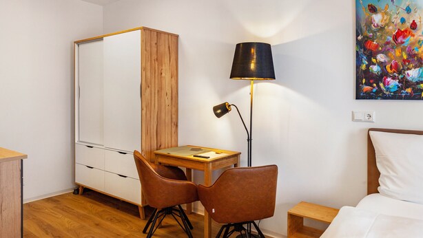 A hotel room with a wooden wardrobe, a desk with a lamp, two chairs, a bed, and a colorful painting on the wall.