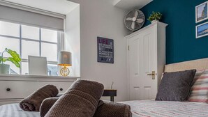 Een net opgemaakt bed met opgerolde handdoeken, een bedlamp, een wandklok, een lijst met een inspirerend citaat, een ventilator en een raam met uitzicht.
