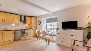 Een moderne keuken met houten kasten, een eethoek met witte stoelen, een televisie op een witte kast en een groot raam.