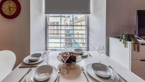 Een eetgedeelte met een tafel voor twee, een raam met uitzicht op gebouwen en een klok aan de muur.