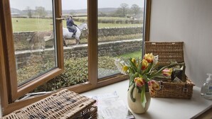 Ein Fenster mit Blick auf eine ländliche Landschaft, ein Korbwägen, eine Vase mit Blumen und eine Flasche Handdesinfektionsmittel auf einem Tisch.