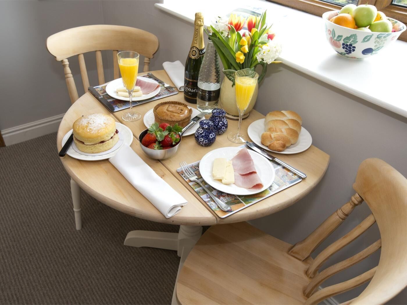 Una mesa de desayuno con una botella de champán, un bol de fruta, un vaso de zumo de naranja, un plato de jamón y queso, un muffin, un croissant y una cesta de fresas.