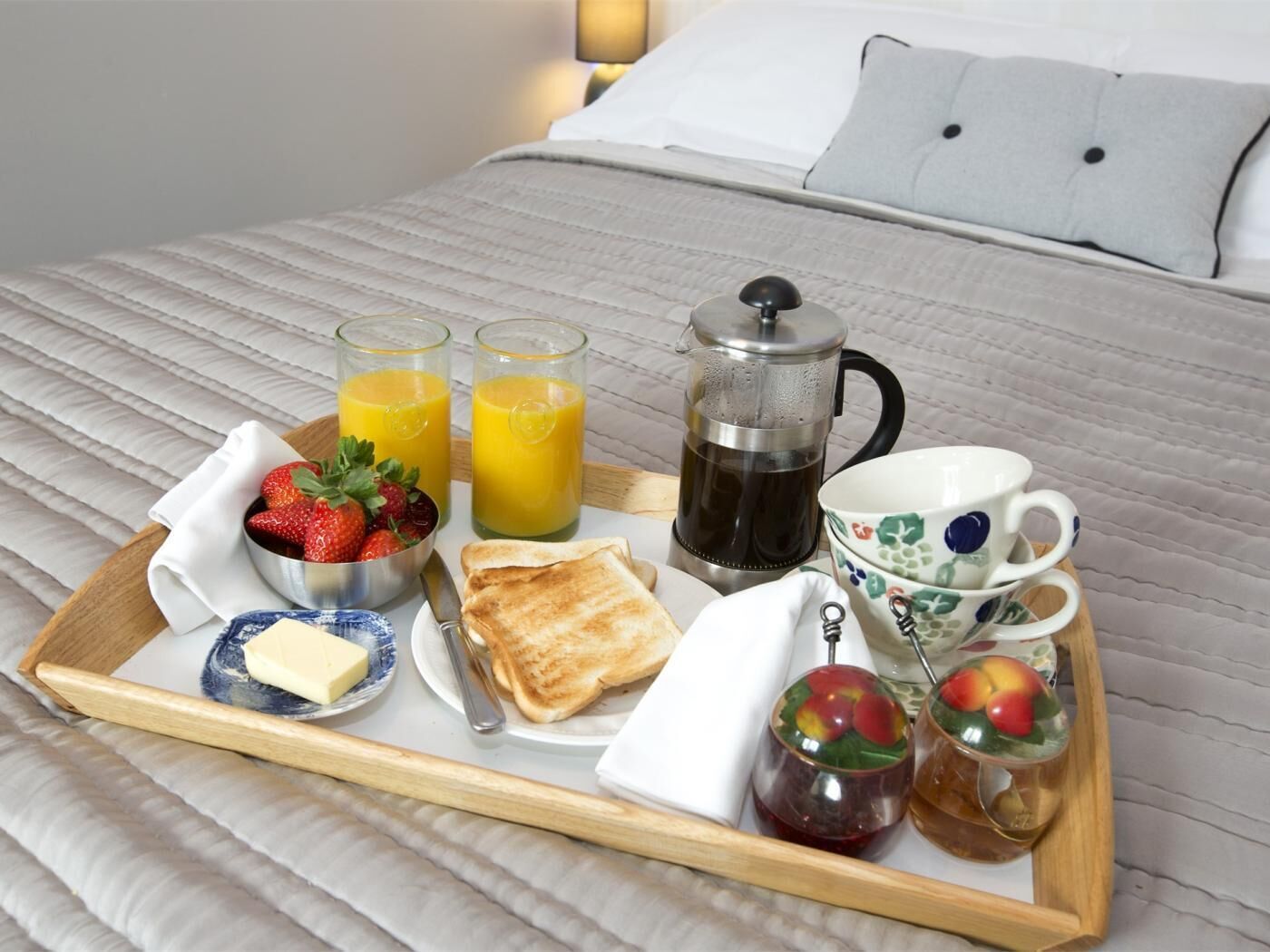 A breakfast tray with toast, fruit, juice, coffee, and a small dish of butter on a bed.