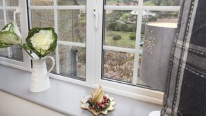Et vindue med udsigt over et landligt landskab, en hvid vase med blomster og en lille pynteting på vindueskarmen.
