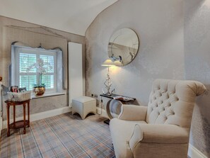A room with a plaid floor, a tufted armchair, a round mirror, a lamp, and a window with an orchid plant. - 3 Bed in Leyburn (oc-g0133) (Preston-under-Scar)