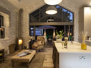A cozy living room with a fireplace, comfortable seating, a dining area with a bar, and a large window overlooking the garden. - 3 Bed in Leyburn (oc-g0133) (Preston-under-Scar)