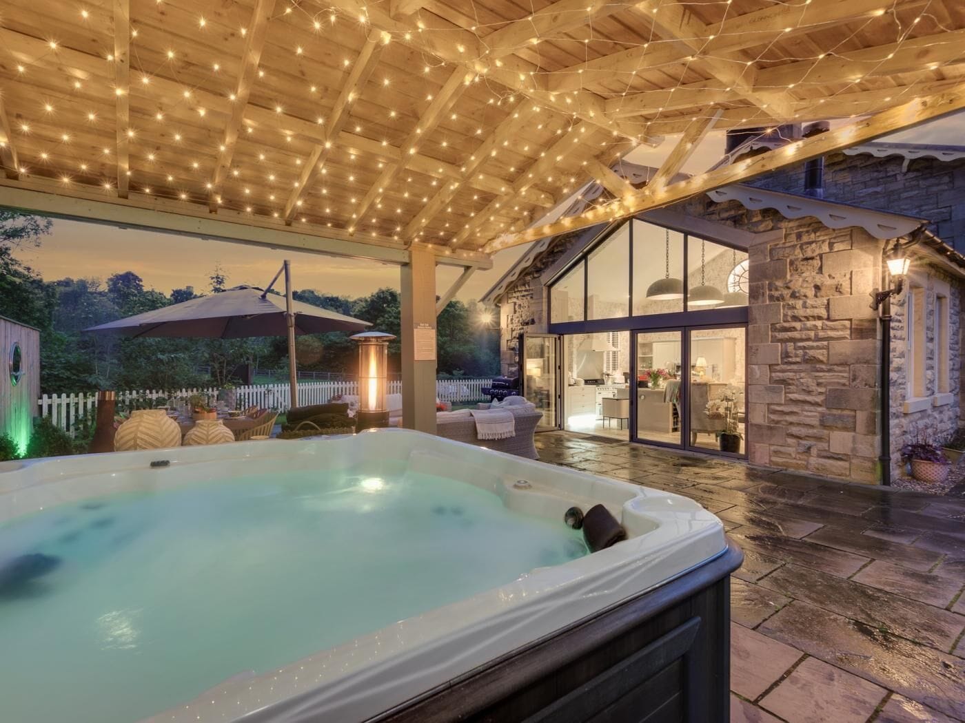 A hot tub under a wooden pergola with string lights, a stone building with glass doors, and a white picket fence in the background.