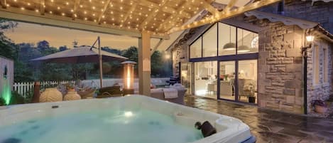 A hot tub under a wooden pergola with string lights, a stone building with glass doors, and a white picket fence in the background.