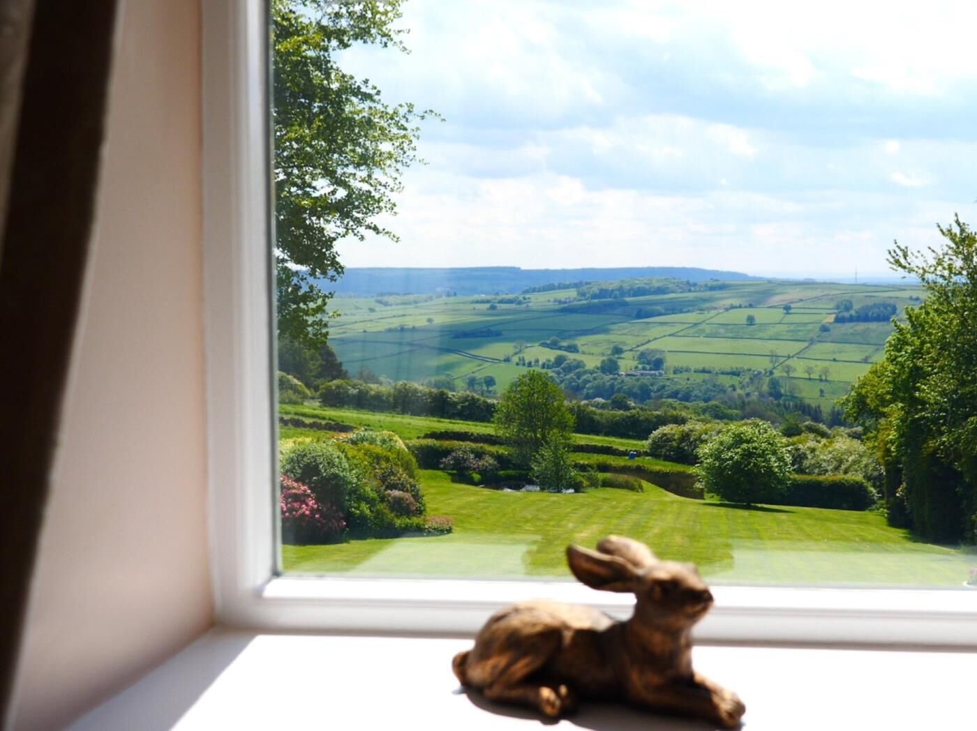 A window with a view of a green landscape, a rabbit figurine on the sill, and a clear sky.