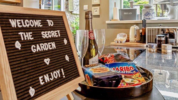 Un angolo cottura con un cartello che recita 'WELCOME TO THE SECRET GARDEN ENJOY!' e una bottiglia di vino sul bancone.