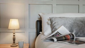 Een net opgemaakt bed met een grijs kussen, een tijdschrift en een kopje op een houten nachtkastje naast een lamp.