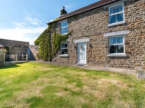 A stone house with a white door and windows, a grassy yard, and a stone wall. - 2 Bed in Bishop Auckland (oc-82620) (Cockfield)