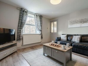 Un salon avec un téléviseur, un canapé, une table basse avec des tasses et une assiette de muffins, une fenêtre avec des rideaux et un radiateur.