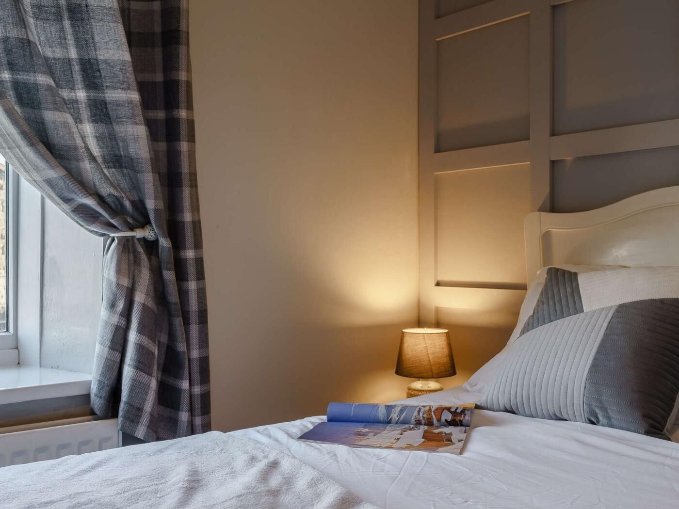A neatly made bed with a pillow and a book, a bedside lamp, and a window with curtains.