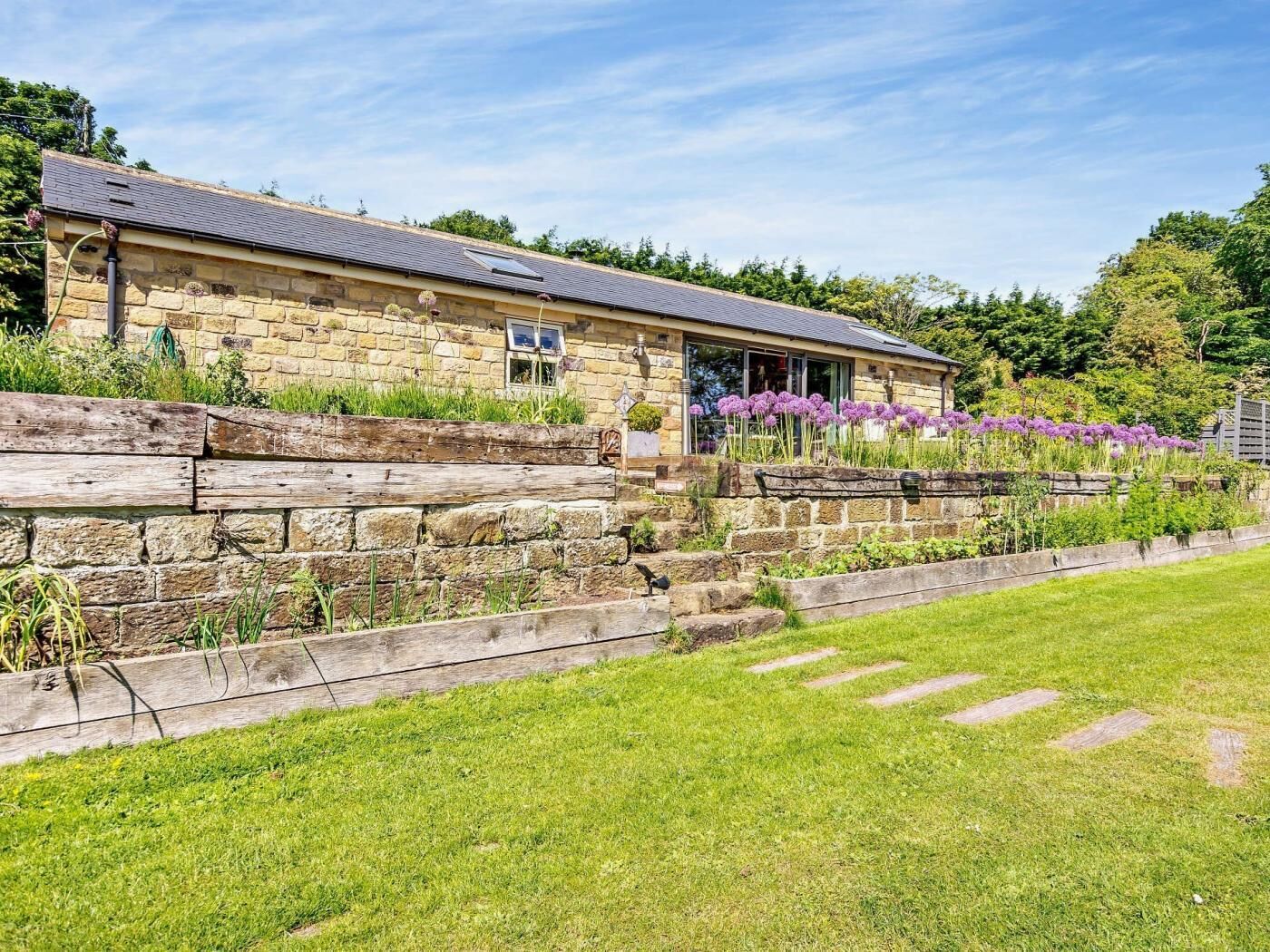 A stone building with a sloped roof, a wooden fence, and a grassy area with purple flowers.