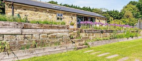 A stone building with a sloped roof, a wooden fence, and a grassy area with purple flowers.