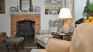 Una sala de estar acogedora con una chimenea de ladrillo, un sillón escocés, una mesa redonda con una lámpara y una estantería llena de objetos decorativos.