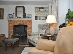 A cozy living room with a brick fireplace, a plaid armchair, a round table with a lamp, and a bookshelf filled with decorative items. - 2 Bed in Chipping Norton (oc-73904) (Swerford)