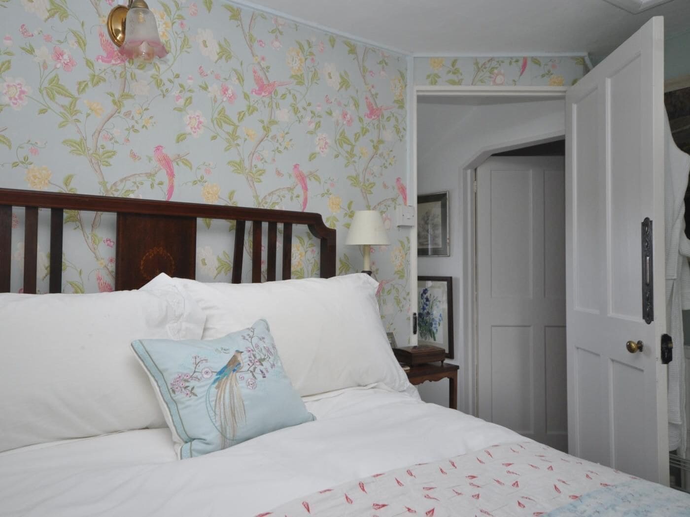 Une chambre à coucher avec un papier peint à motifs floraux, une tête de lit en bois, un lit blanc avec un oreiller bleu, une table de chevet avec une lampe et une porte donnant sur une autre pièce.