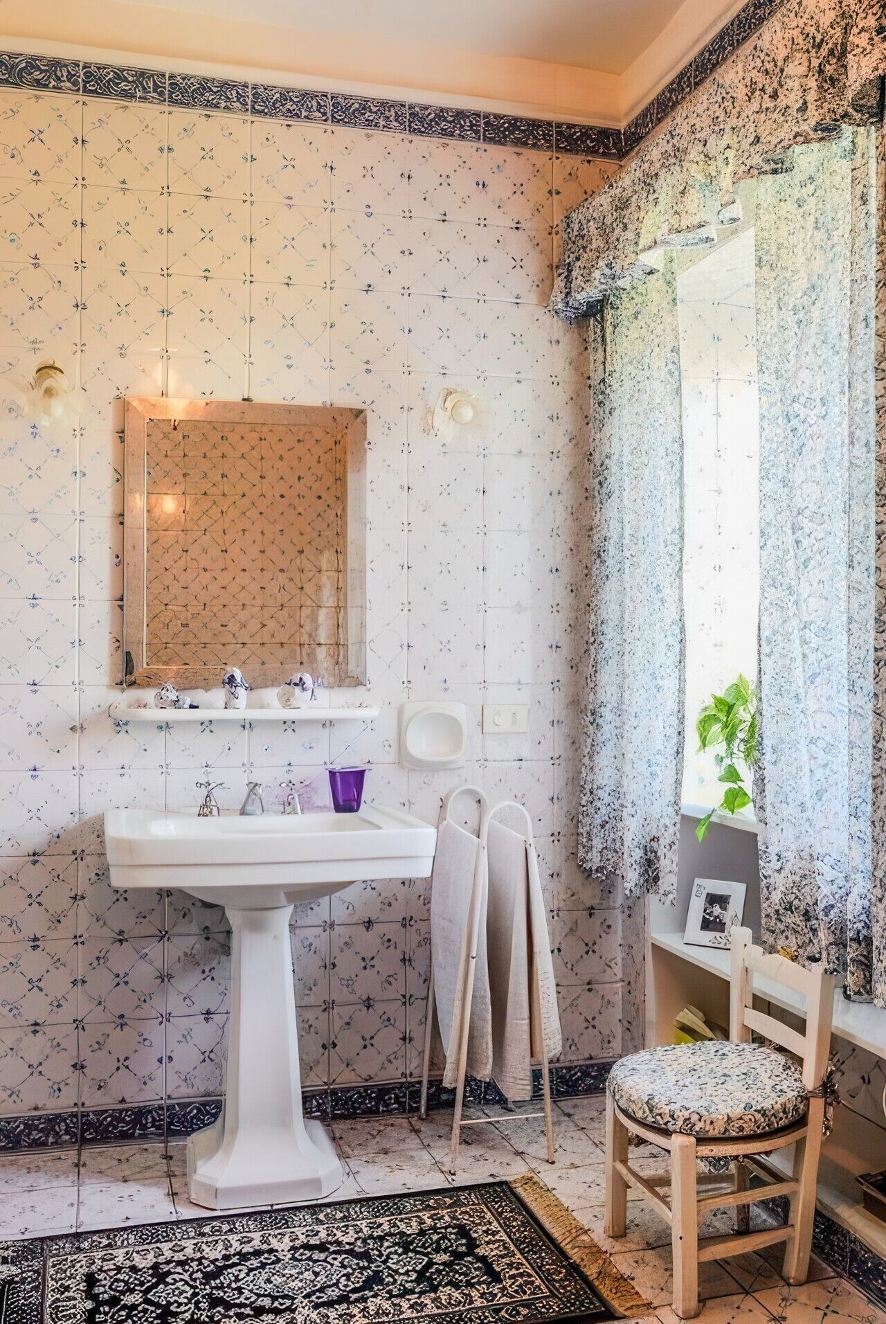 Un baño con un lavamanos blanco de pedestal, un espejo encima, un perchero, una silla de madera con cojín estampado, y un suelo con diseño.
