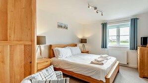 A bedroom with a wooden bed, two bedside lamps, a plaid chair, a TV, and a window with curtains. - 3 Bed in Woolacombe (oc-74255) (Woolacombe)