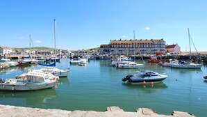 A marina with various boats docked, a building with multiple windows, and a clear sky. - 4 Bed in Bridport (oc-dc083) (West Bay)