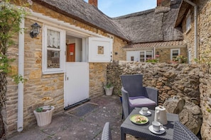 A stone building with a thatched roof, a door, a wicker chair, a table with a plate of food, and a potted plant. - 2 Bed in Bridport (oc-dc148) (Burton Bradstock)