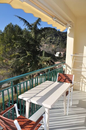 Un balcon avec une table et des chaises, un parasol jaune et une vue sur les arbres et les bâtiments.