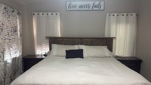 Ein ordentlich bezogenes Bett mit weißer Bettwäsche und einem dunklen Kopfkissen, flankiert von zwei Nachttischen mit Lampen, in einem Zimmer mit Holz-Kopfteil und einem motivierenden Zitat an der Wand.