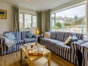 A living room with two striped sofas, a wooden coffee table, and a window with a view of houses and greenery. - 3 Bed in Bridport (oc-dc117) (West Bay)