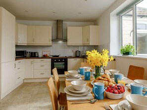 A modern kitchen with white cabinets, a stove, and a dining table set with blue cups, plates, and a vase of yellow flowers. - 3 Bed in Bridport (oc-dc117) (West Bay)