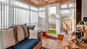 Ein gemütliches Zimmer mit einem Korbsessel, einem Esstisch mit Kaffeemaschine und Blick auf den Garten durch große Fenster.