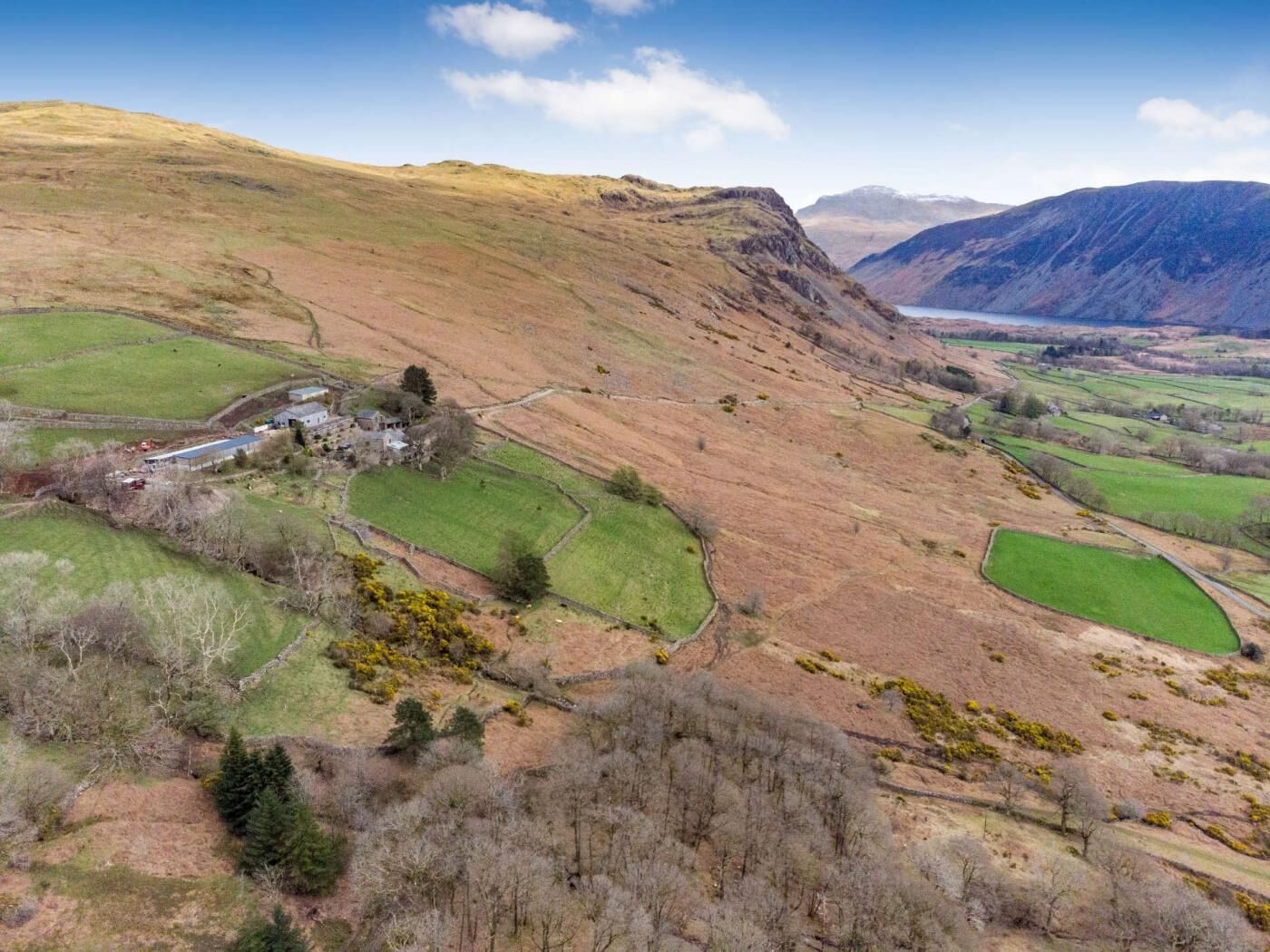 Paesaggio rurale con dolci colline, un piccolo gruppo di edifici e un lago in lontananza.