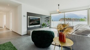 A modern living room with a flat-screen TV, a dark green chair, a round gold table with a vase, and a grey sofa.